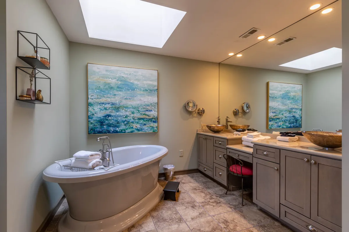 A serene bathroom with a freestanding soaking tub positioned under a window, allowing in abundant natural light. The decor includes elegant artwork and a spacious vanity area, emphasizing tranquility and relaxation.