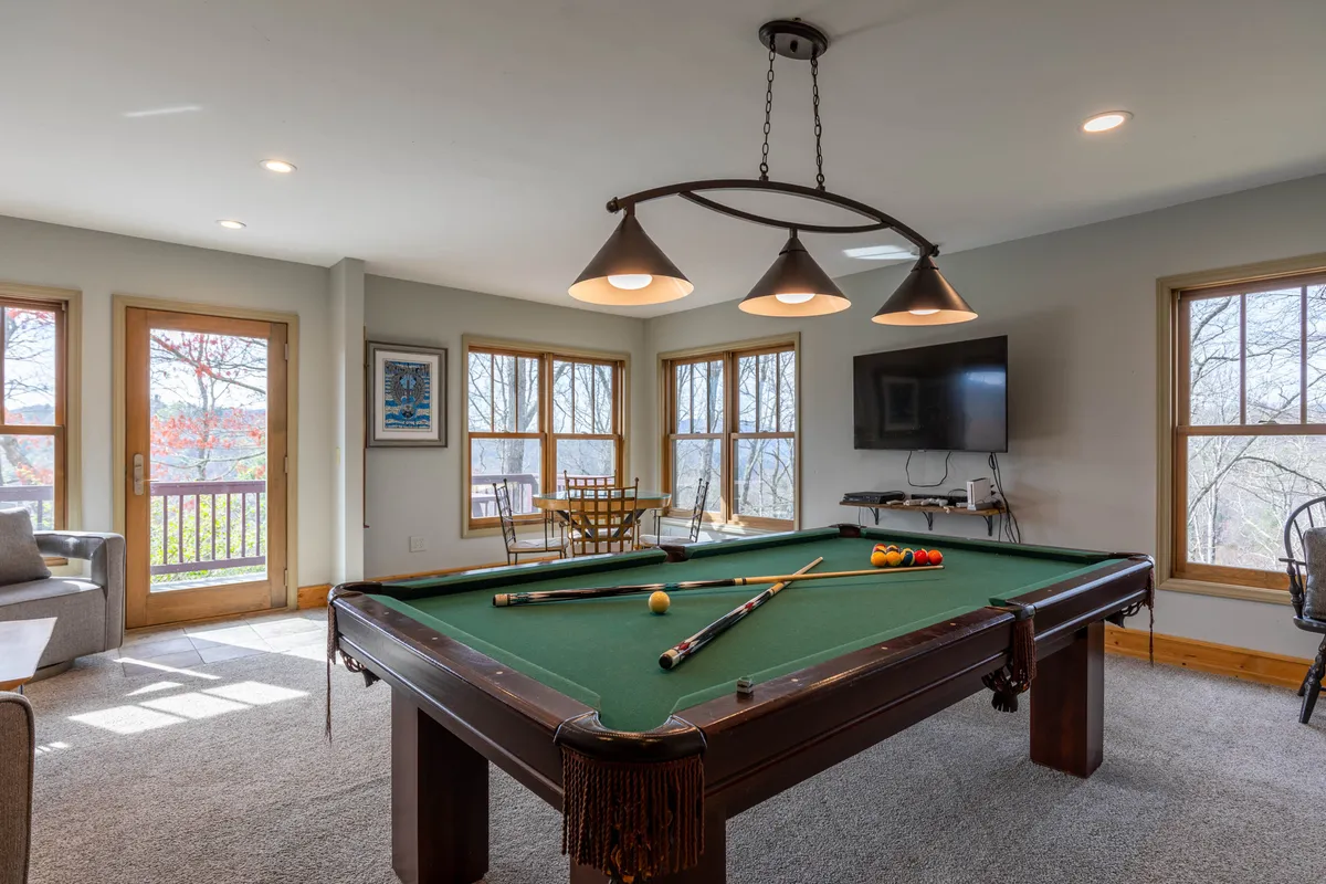 This vibrant interior scene centers on a pool table set beneath overhead lighting, with a wall-mounted TV and expansive windows offering views and natural light. The space is casual and inviting, crafted for socializing and friendly competition.
