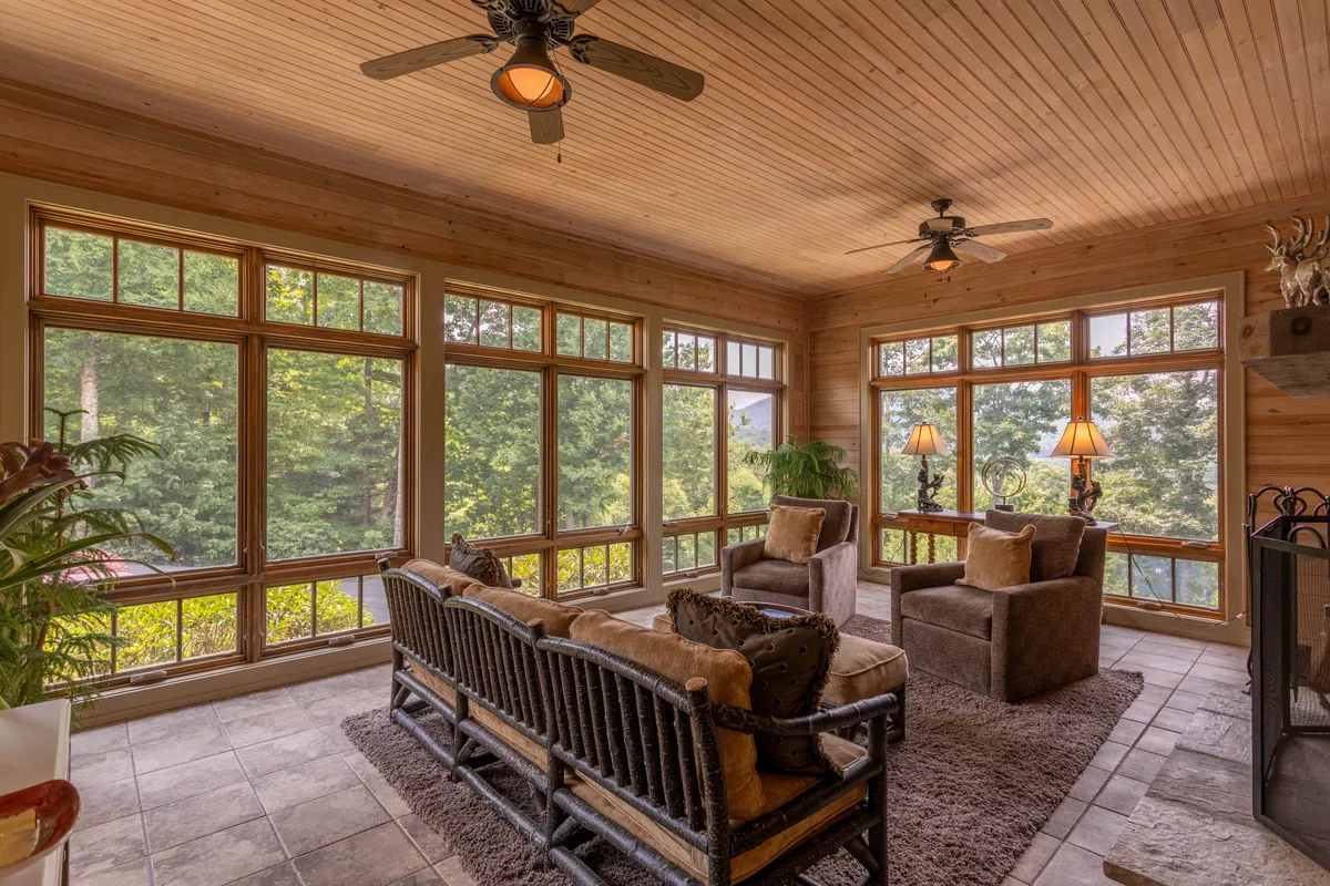A tranquil sunroom with large windows offering panoramic views of the surrounding nature. Plush, comfortable seating is arranged to take advantage of the views and natural light, with decorative lamps and a textured area rug adding warmth and coziness to the space.