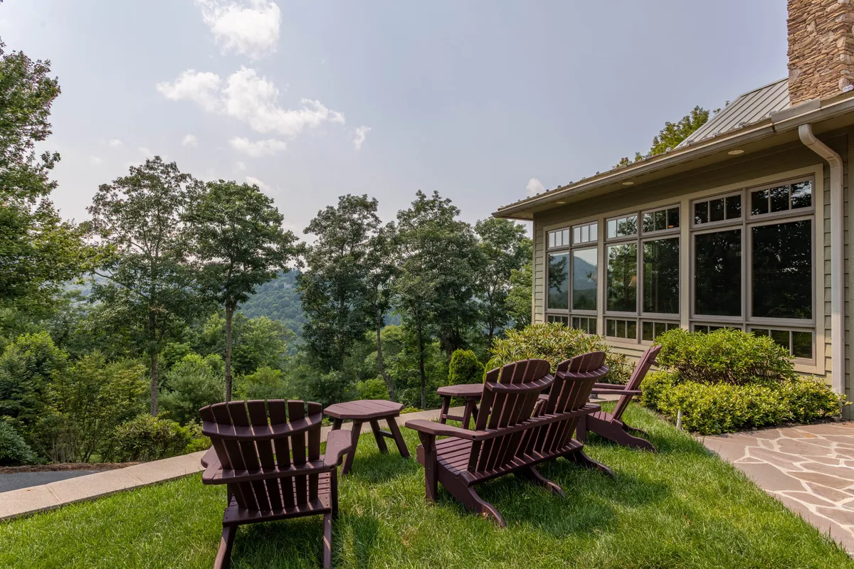 A serene stone patio featuring a pair of striking purple Adirondack chairs, set against a lush green lawn and distant rolling mountains. The area is surrounded by natural landscaping and offers open sky views, evoking a sense of peaceful retreat and connection to the outdoors.