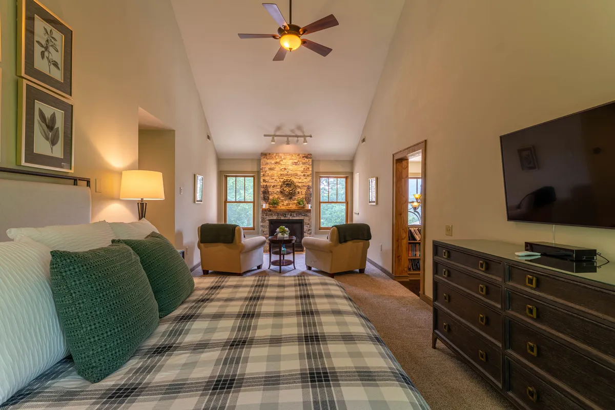 A spacious bedroom featuring a cozy bed with plaid bedding, a warm stone fireplace, a seating area with two armchairs, a television, and decorative artwork. Natural light fills the room, creating an inviting atmosphere ideal for relaxation.