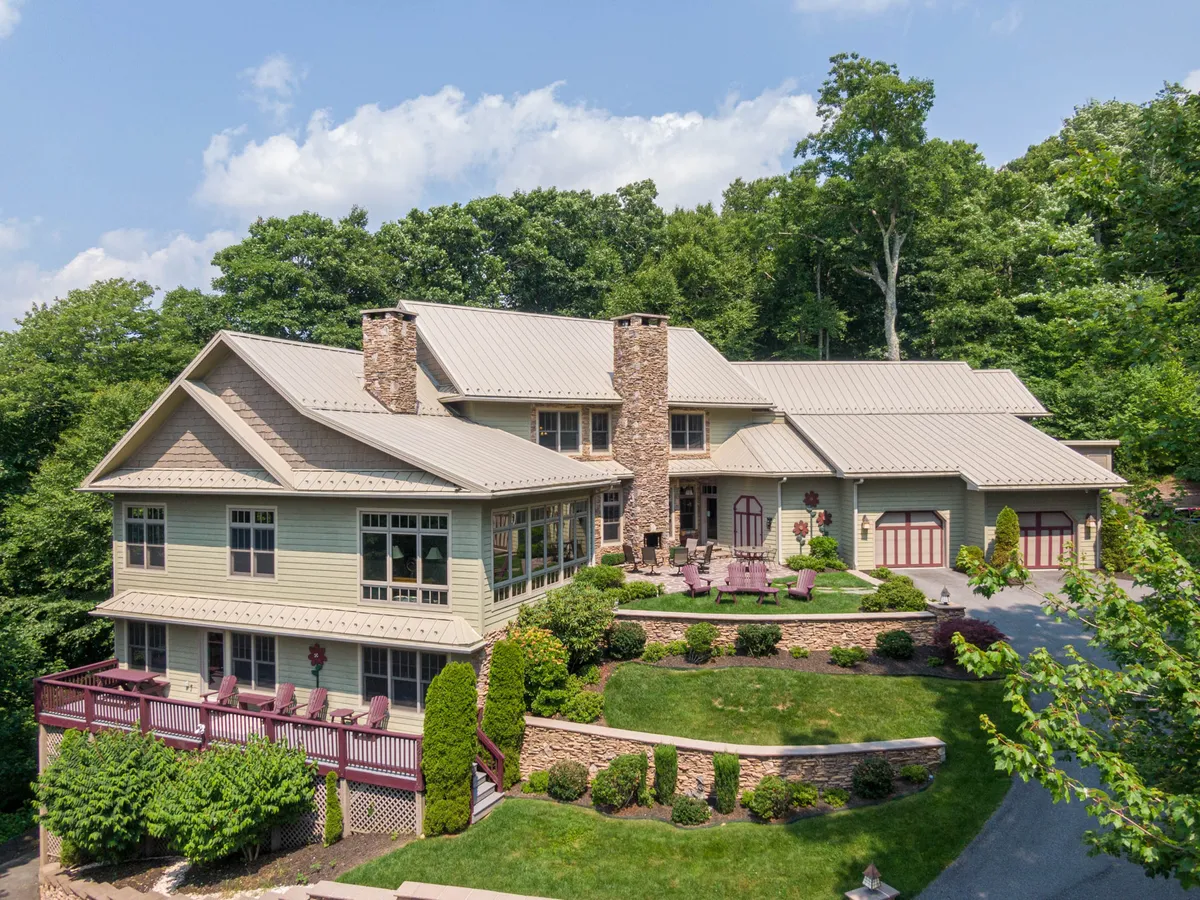 The hero image showcases the grand exterior of a luxury mountain home, featuring a striking stone chimney, a spacious deck perfect for gatherings, beautifully landscaped gardens, and multiple roof lines that add architectural interest. The inviting scene is set against the serene backdrop of Blowing Rock's rolling hills, with golden sunlight casting a warm glow over the outdoor spaces, hinting at relaxing moments and stunning sunset views.