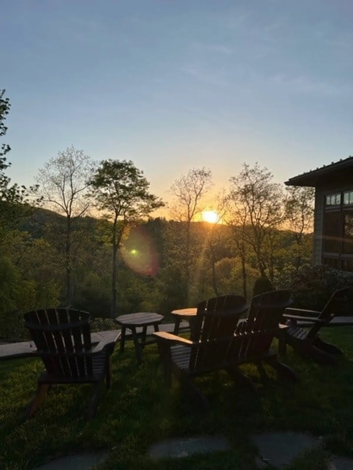 This peaceful exterior scene features Adirondack chairs arranged to face a stunning mountain sunset, surrounded by trees and lush grass. The tranquil mood is ideal for sharing a glass of wine, reflecting, or unwinding at the end of the day.